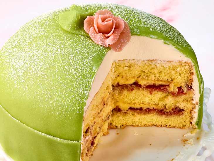 Layered cake with green marzipan icing, pink rose decoration, and visible layers of sponge, jam, and cream.