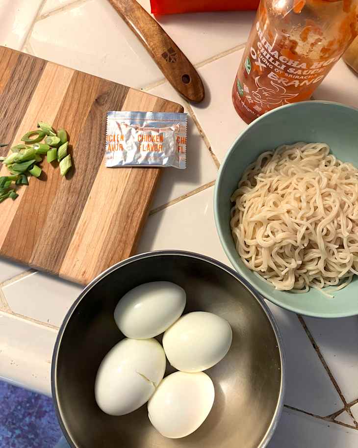 Bowl with four boiled eggs, a packet of chicken flavoring, green onions on a cutting board, and cooked noodles in a bowl.