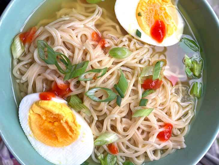 Bowl of noodles in broth topped with sliced green onions, boiled egg, and chili sauce.