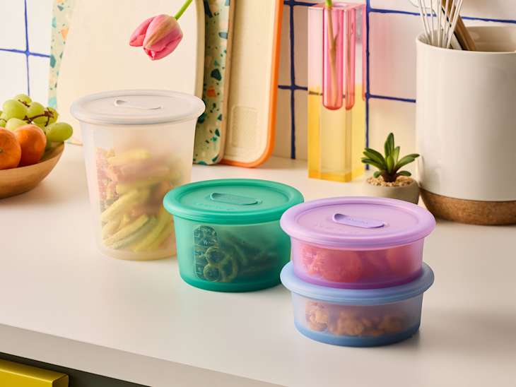 Colorful food storage containers on a kitchen counter with fresh fruits, a plant, and utensils in the background.