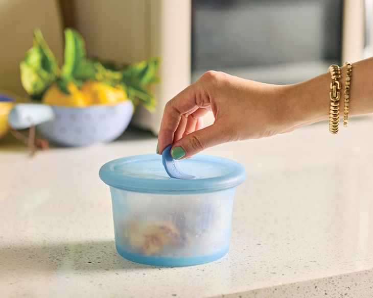 A hand with a gold bracelet lifts a blue silicone lid from a clear container on a kitchen countertop.